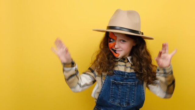Scary girl child with Halloween makeup mask wears hat and shirt, dance gesticulating hands fool around have fun enjoy celebrate isolated on yellow color background in studio. Party holiday concept