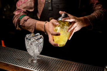 crystal goblet with ice cubes on bar and male hands holding glassy shaker with cocktail