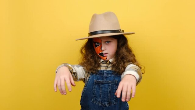 Portrait of scary bright alive fun little girl with Halloween makeup mask wears hat and shirt, hands raised, looking at camera, posing isolated on yellow color background in studio. Holiday concept