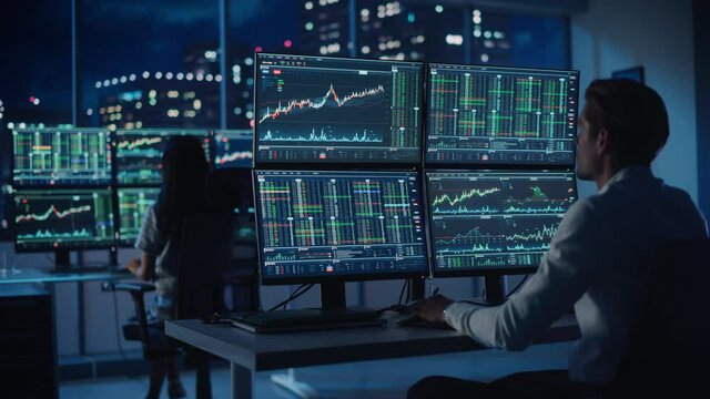 Financial Analyst Working on a Computer with Multi-Monitor Workstation with Real-Time Stocks, Commodities and Foreign Exchange Charts. Businessman Works in Investment Bank City Office at Night.