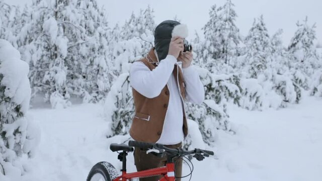 Male traveler standing near fat bike and taking photos on retro camera in snowy winter forest 