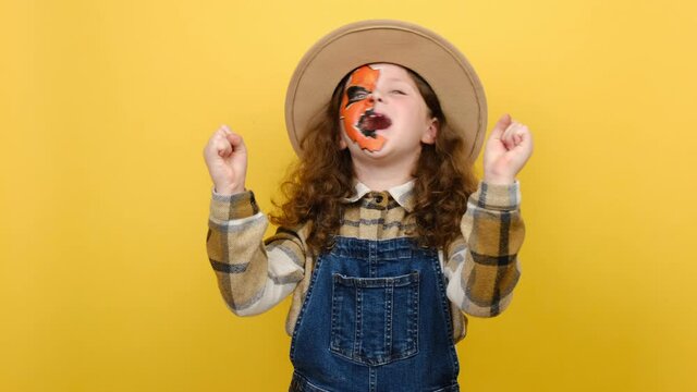 Portrait of scary bright vivid little girl child with Halloween makeup mask wears brown hat and shirt look camera surprised celebrating clenching fists isolated on yellow color background in studio