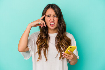 Young caucasian woman holding a mobile phone isolated on blue background showing a disappointment gesture with forefinger.