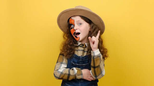 Portrait of insighted smart little girl child with Halloween makeup mask wears brown hat and shirt looks around thinks comes up with ideas raised finger, posing isolated on yellow background studio