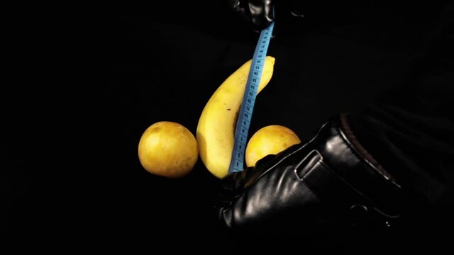 A pair of hands in black leather gloves measuring a ripe yellow banana with a tailor's tape (also, two plums are there). Visual metaphor: checking the size of a specificl human anatomical part.
