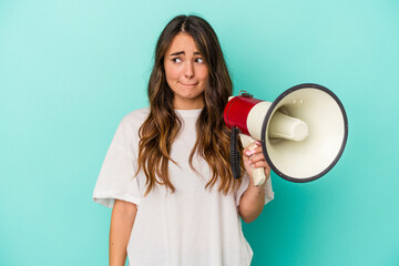 Young caucasian woman holding a megaphone isolated on blue background confused, feels doubtful and unsure.