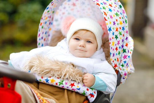 Cute little beautiful baby girl sitting in the pram or stroller on autumn day. Happy healthy child going for a walk on fresh air in warm clothes. Baby with in colorful clothes and hat with bobbles