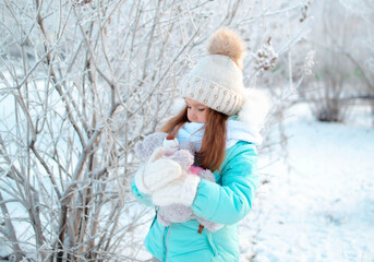 A small, charming girl in winter in a snowy park holds a teddy bear. Background of a winter forest landscape