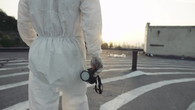 Unrecognizable worker in protective suit walks with mask and respirator in hands
