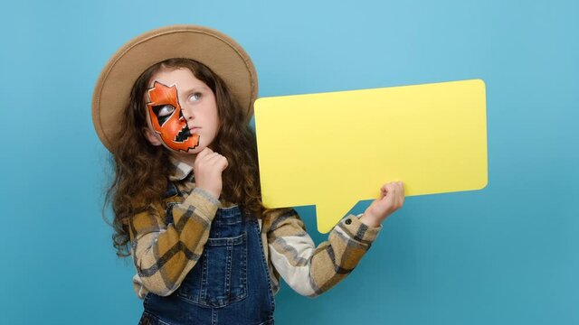 Thoughtful little girl with Halloween makeup mask holding an empty speech bubble, ponders on decision, thinking about something, isolated over blue background with copy space. Hmm something strange