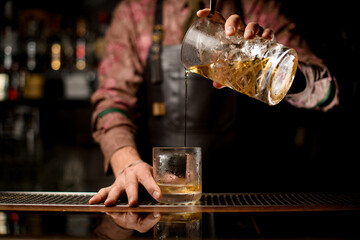 Fototapeta na wymiar hand of barman holds crystal mixing cup with drink and pour it into a glass at the bar
