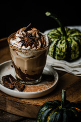 Pumpkin pie spice mocha latte with whipped cream in glass with dark chocolate on plate on wooden tray with pumpkins on dark background .