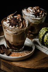 Two glasses of Pumpkin pie spice mocha latte with whipped cream and dark chocolate on plate on wooden tray with pumpkins on dark background .