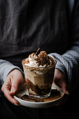 Woman in sweater and dark apron holding plate with  glass of Pumpkin pie spice mocha latte with whipped cream  and dark chocolate  on black background .