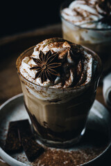 Pumpkin pie spice mocha latte with whipped cream in glass and dark chocolate on plate on wooden table on dark background .