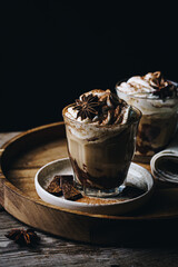 Two glasses of Pumpkin pie spice mocha latte with whipped cream and dark chocolate on plate on wooden tray on dark background .