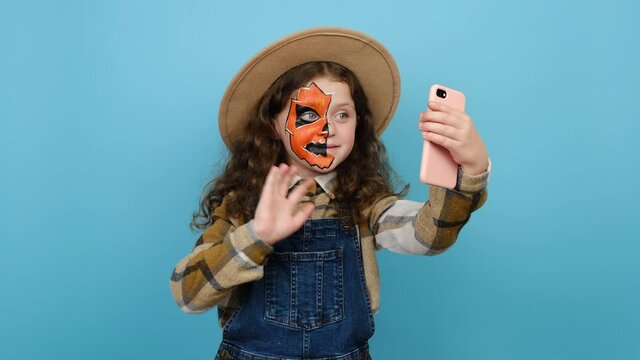 Portrait of happy cute fun little girl kid with Halloween makeup mask wears hat get video call using mobile cell phone talk greet with hand, posing isolated over blue color background in studio