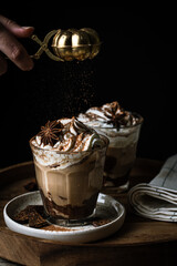 Sprinkling Pumpkin pie spice on mocha latte with whipped cream in glass with dark chocolate on plate on wooden tray on dark background .