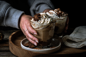 Woman in sweater holding two glasses of Pumpkin pie spice mocha latte with whipped cream  and dark chocolate  on wooden tray black background .