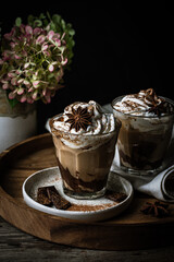 Two glasses of Pumpkin pie spice mocha latte with whipped cream and dark chocolate on plate on wooden tray on dark background .