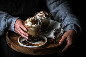 Woman in sweater and dark apron holding wooden tray with  two glasses of Pumpkin pie spice mocha...
