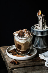 Pumpkin pie spice mocha latte with whipped cream in glass, dark chocolate on plate and natural wax candle on wooden table on dark background .