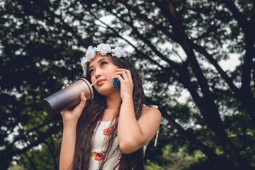 A teenage lady wearing a flower crown calling her friend while taking a sip from her stylish...