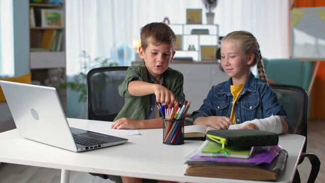 kids learn online, cute boy pupil studying with girl with a plaster cast on the arm while sitting at home at table with computer laptop