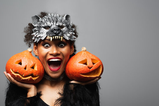 astonished african american woman in wolf halloween mask holding carved pumpkins isolated on grey