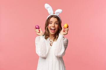 Holidays, spring and party concept. Portrait of excited charming young woman celebrating Easter in rabbit ears and white party dress, dancing with two painted eggs, pink background