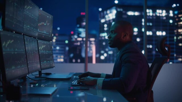 Financial Analyst Working on Computer with Multi-Monitor Workstation with Real-Time Stocks, Commodities and Exchange Market Charts. African American Businessman Works in Investment Bank Late at Night.