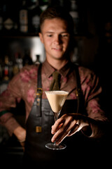 hand of male bartender gently holds martini glass with cocktail. Blurred bartender in background