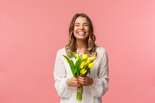 Holidays, beauty and spring concept. Portrait of happy excited charming blond girl receive flowers, buying yellow tulips herself, smiling and laughing joyfully, stand pink background