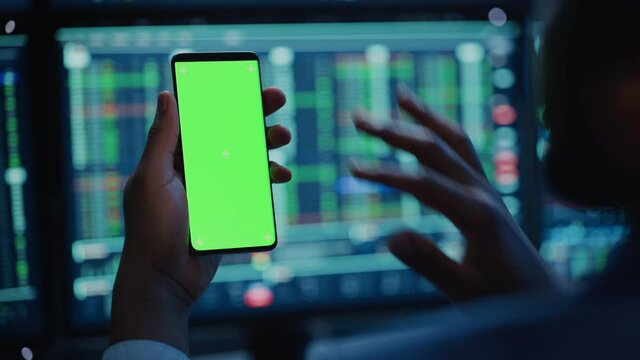 Financial Analyst Using Smartphone with Green Screen Chroma Key Mock Up Template and Working on Multi-Monitor Workstation with Stocks Charts. Businessman Works in Investment Bank in the Evening.