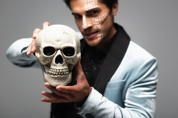 blurred man in halloween makeup showing scary skull while looking at camera isolated on grey