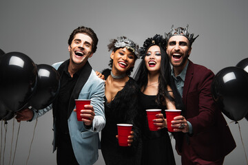 cheerful multicultural friends with plastic cups smiling at camera near black balloons on halloween party isolated on grey