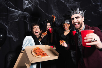 excited man in halloween makeup showing pizza near happy multiethnic friends on black