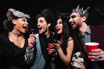 joyful multiethnic friends in spooky costumes singing karaoke on halloween party on black