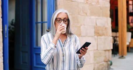 Enjoyment. Motion camera view of the senior woman looking at the smartphone and drinking coffee while waiting for somebody at the street. People lifestyle concept