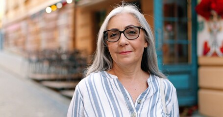 Portrait view of the happy senior wrinkled woman wearing glasses looking at the camera with pleasure smile while walking alone at the city. Elderly people concept