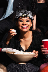excited african american woman in wolf mask eating popcorn during halloween party on black