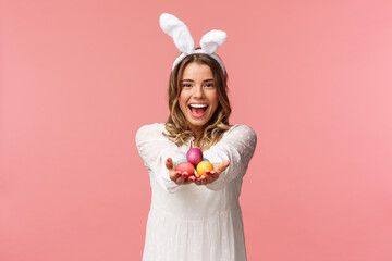 Holidays, spring and party concept. Portrait of lovely, romantic young blond woman in rabbit ears and white dress, giving you painted easter eggs as celebrating orthodox holiday, pink background