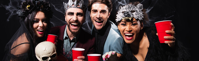 excited multiethnic friends with plastic cups laughing at camera during halloween party on black,...