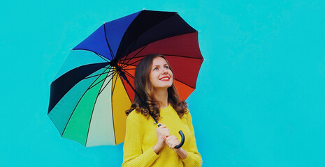 Autumn portrait of happy cheerful smiling young woman with colorful umbrella wearing an yellow knitted sweater on blue background