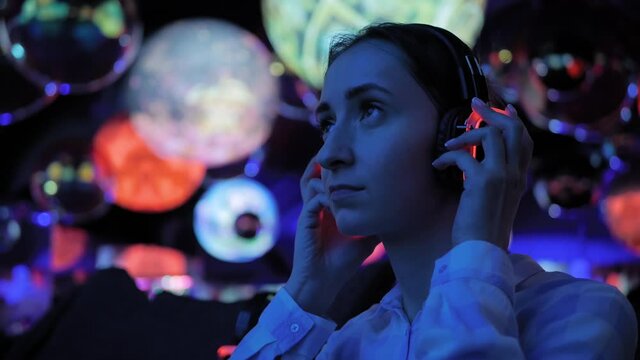 Woman wearing wireless black headphones and looking around in dark room of interactive museum or exhibition with colorful illumination. Immersive, futuristic, entertainment concept