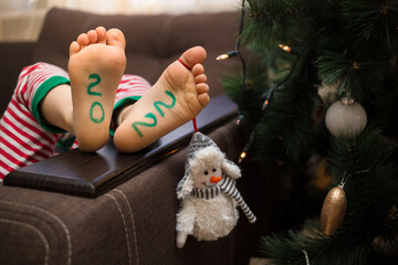 barefoot kid in striped pants lies on the sofa near the Christmas tree. Drawn numbers 2022 on the feet. Cozy, cheerful, festive atmosphere. Waiting for the holiday. Preparing for the New Year