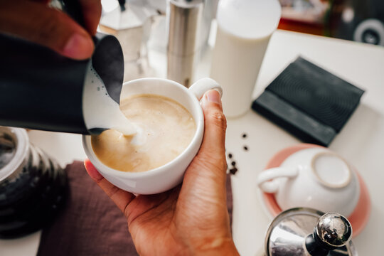 barista's hand making hot coffee mugs, latte art and pitcher made of milk froth, and there are coffee-making accessories on the white table. Close up