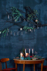 Elegant Christmas table setting with cozy candles, plates, pomegranate and branch of fir tree. Vintage blue wall copy space with branch and bulbs. Cozy home concept