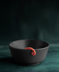 Curled octopus leg coiling out of grey bowl on plain dark background.