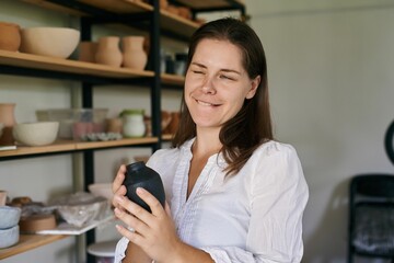Fototapeta na wymiar woman artisan ceramist stands against rack handmade clay utensils, holds small black vase